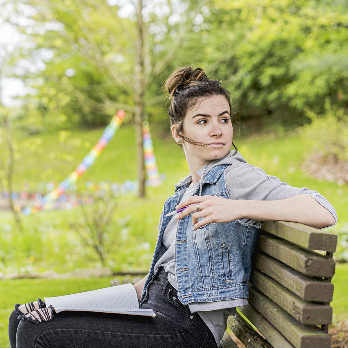 Photo of a young white woman with hair tied up, wearing a jean jacket 和 seated on a bench outside looking behind her into the distance